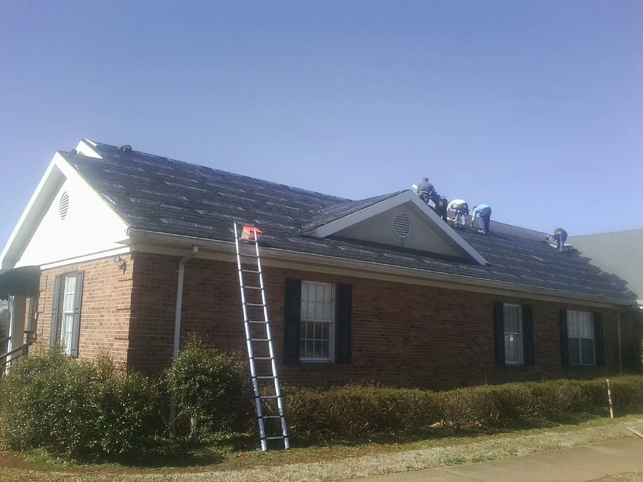 Crew tearing shingles off a brick ranch roof with a ladder set — Emergency Roof Repair work by our crew in Tipton